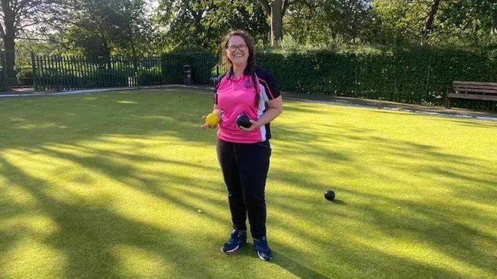 A woman in a pink top and black trousers with dark hair and glasses, standing on a bowling green holding two bowls