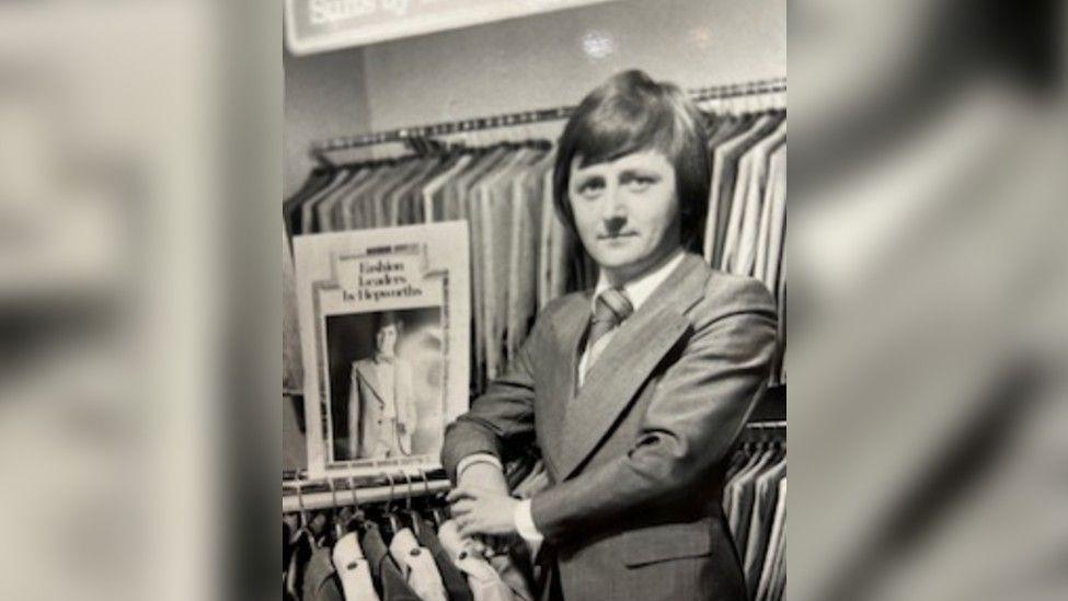 A black and white photo showing a man in a suit stood in a clothing shop surrounded by suit jackets. He is clean shaven and has a mod haircut style. 
