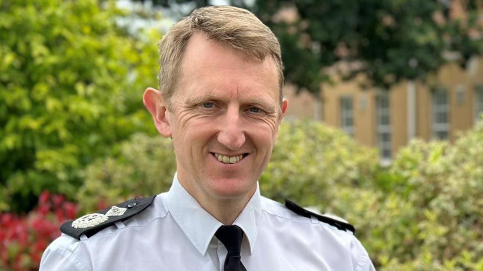 A man smiling at the camera wearing police uniform. He is wearing a white shirt with a black tie with police badges sewn on to black lapels on his shoulders. he is stood outside in front of greenery trees and bushes