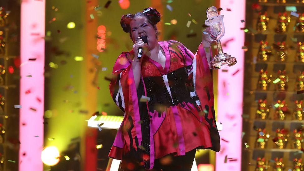 A woman appears with confetti around her as she holds the Eurovision trophy.