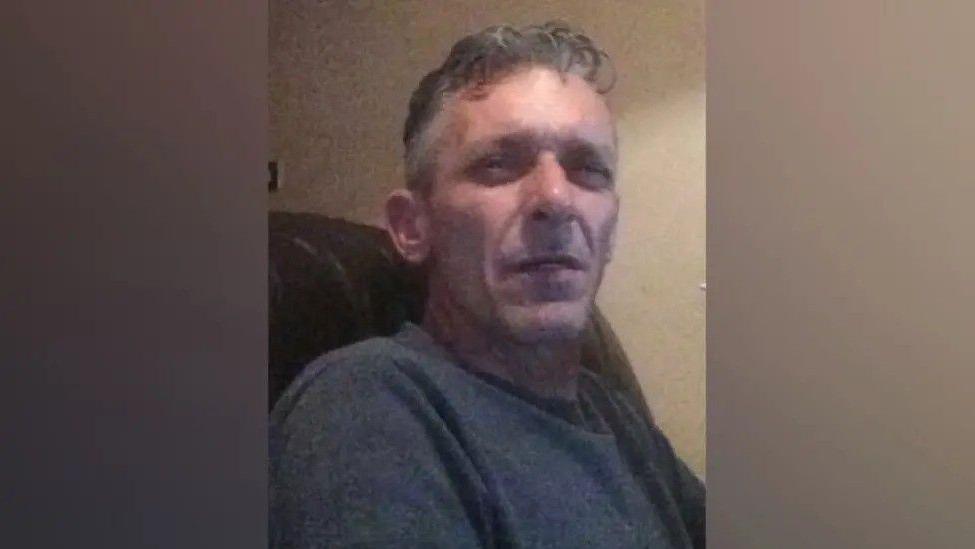 A man in his mid-50s with grey hair. He is sitting in an armchair and squinting at the camera.