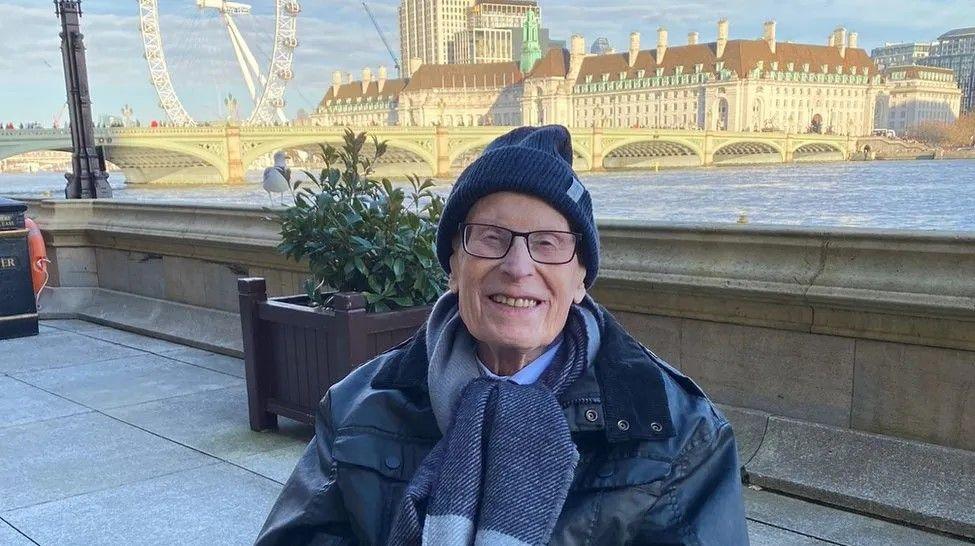 An elderly man in a jacket, scarf, and hat smiles at the camera, on a balcony overlooking the River Thames, the London Eye, and a bridge across the river