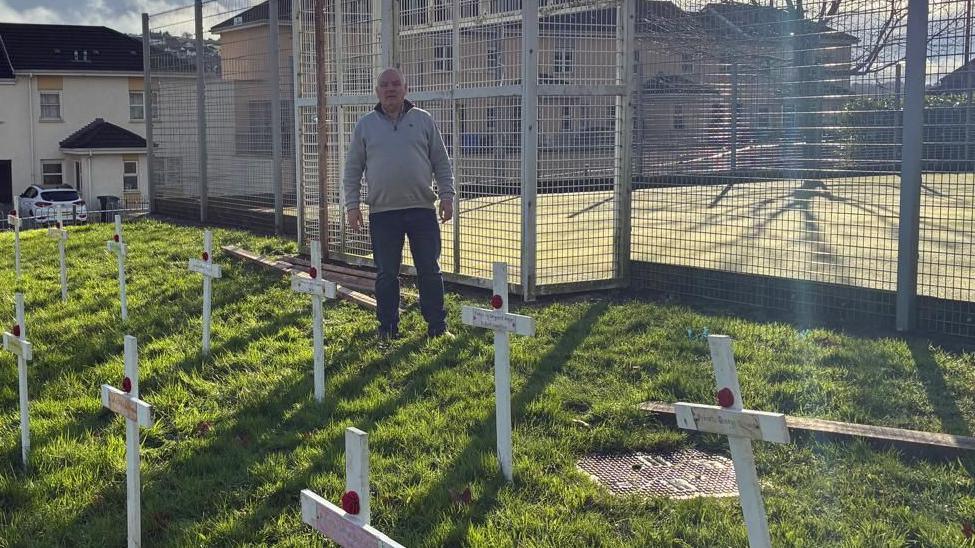 Shows a man standing in a grassy area where white crosses are placed in the ground with poppies and people's names on them.