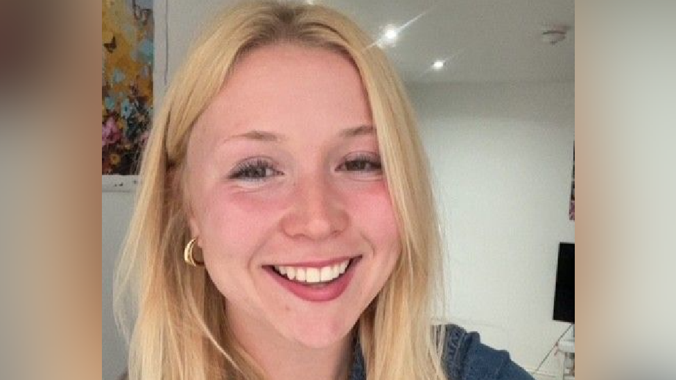 Aneesa is a young woman with long blonde hair worn down. She smiles at the camera and wears a denim top and gold necklace and earrings