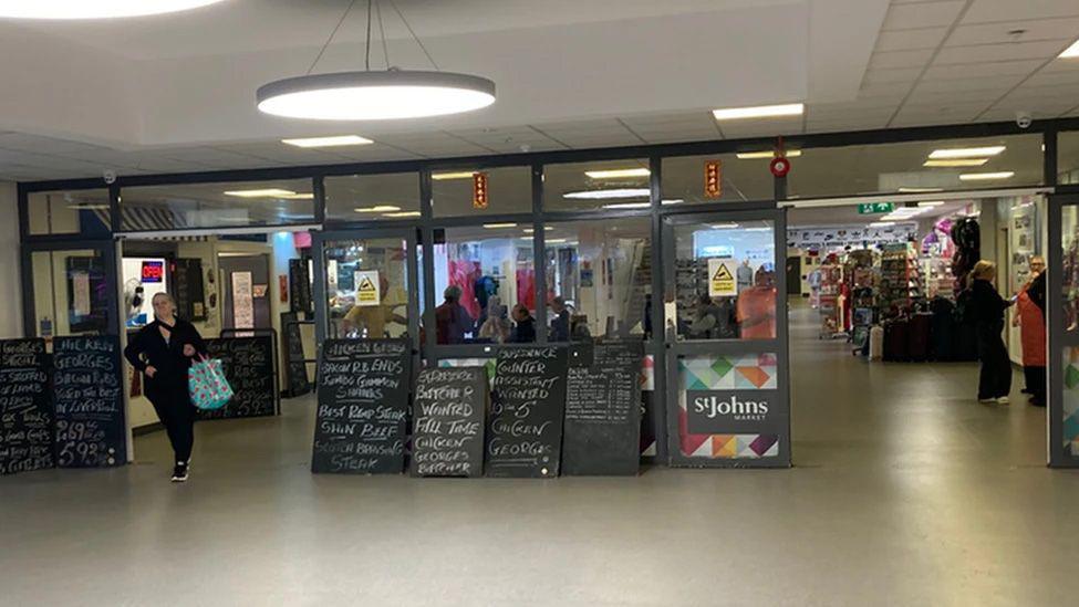 Entrance to St Johns Market when it was open with black boards with writing in chalk and neon lights saying open. A woman in walking through the door with a blue carrier bag and two women are standing in side the market