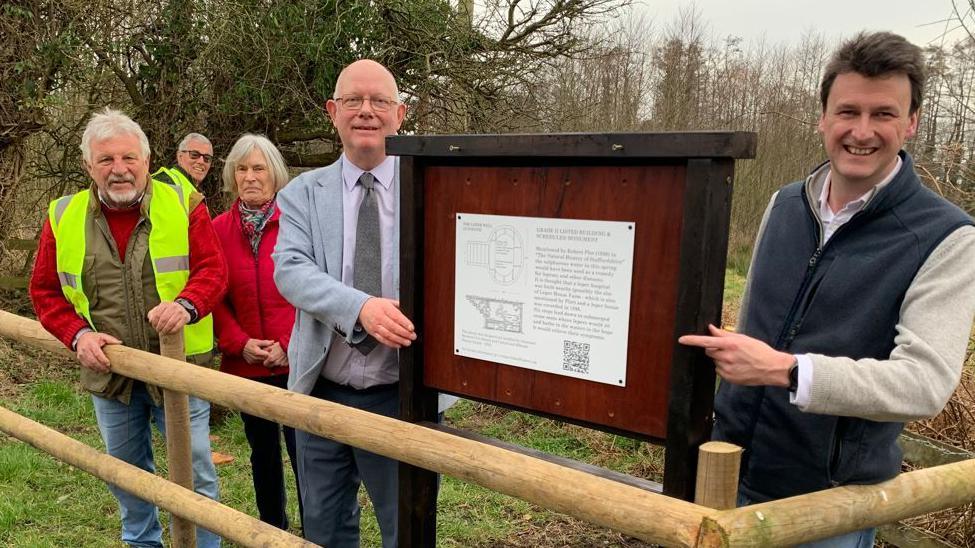 Four men and one woman stood behind a wooden fence in front of some trees, beside a wooden sign post with a white plaque on it