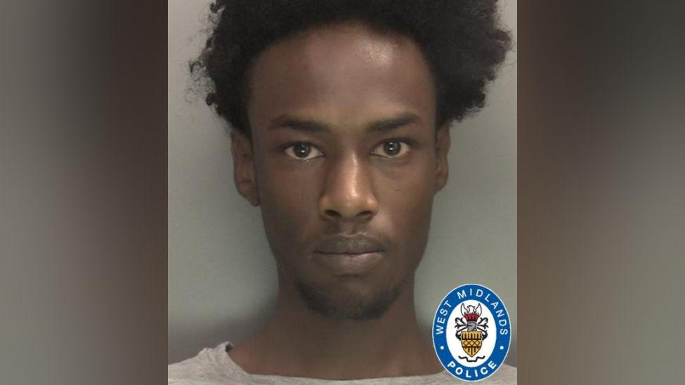 A police mugshot showing a man with curly black hair and brown eyes, wearing a grey T-shirt. The police logo is in the bottom right-hand corner of the photo.