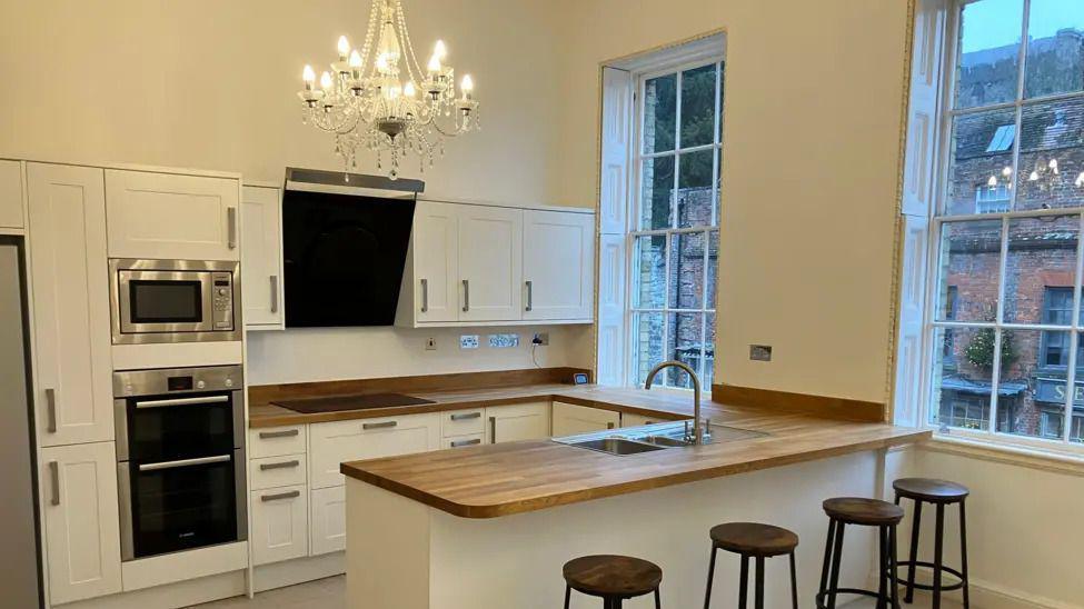 A kitchen in a flat with white cupboards and a light wood breakfast bar. There is a chandelier hanging from the ceiling and two large windows on the right.