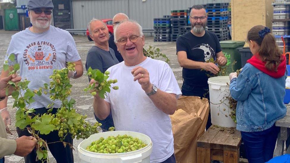 A group of people looking to camera with hops in their hands  with a brewery in the background 