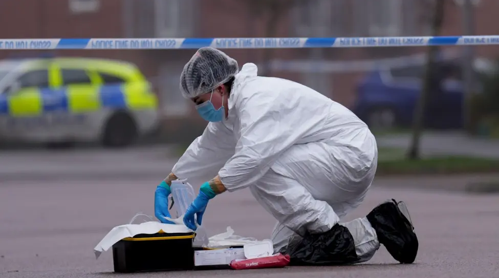 A forensics officer in a white suit, blue gloves, a facemask and a hairnet kneels on the road in front of police tape. There is a case full of equipment in front of them.