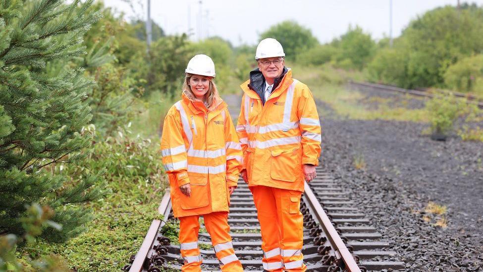 A blonde woman standing next to an older man. Both are wearing orange hi-vis jackets and trousers and hard hats. They're standing on a railway line surrounded by green bushes.