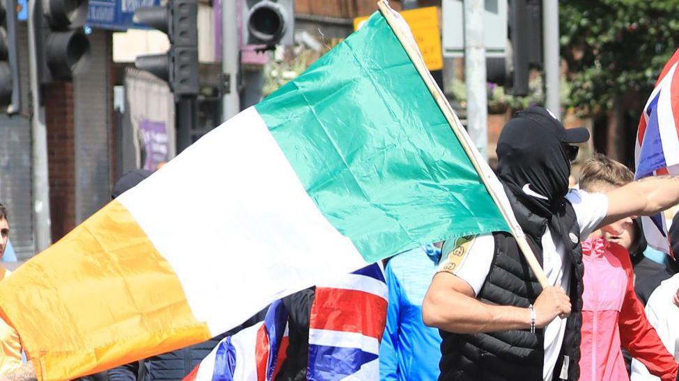 A close up of a man carrying a large Irish tricolour flag at an anti-immigration protest in Belfast on 3 August. Bernard Lavery is wearing a which T-shirt, a black bodywarmer, a black baseball cap and dark glasses and his head is covered with a black garment