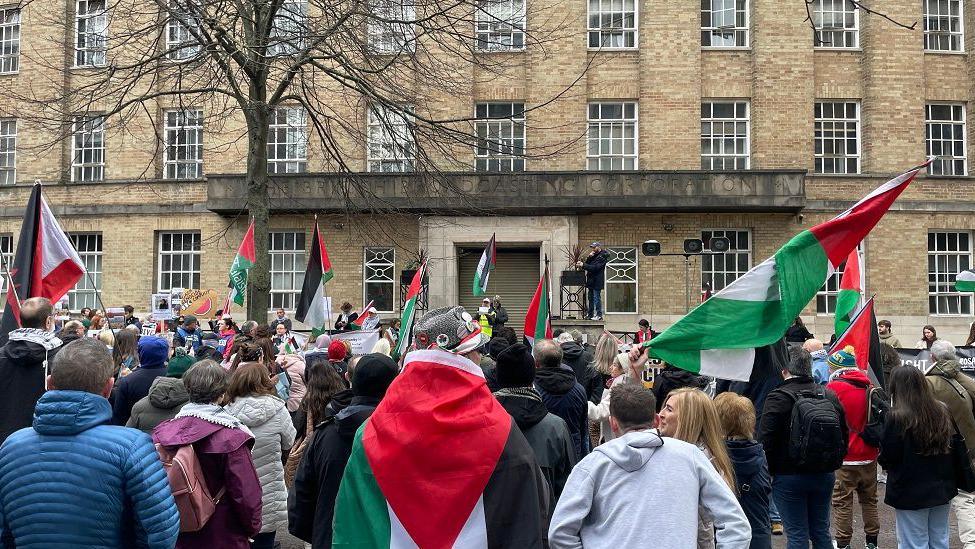 A crowd outside a light stone building holding Palestinian flags. One person is standing up on the wall taking a picture. 