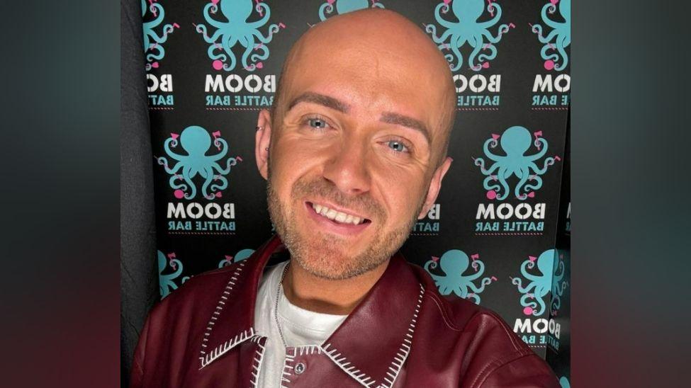 A close-up photo of a man standing against a patterned backdrop. He has a shaved head  blue eyes, and stubble. He is wearing a maroon leather jacket and a white T-shirt. He is smiling.