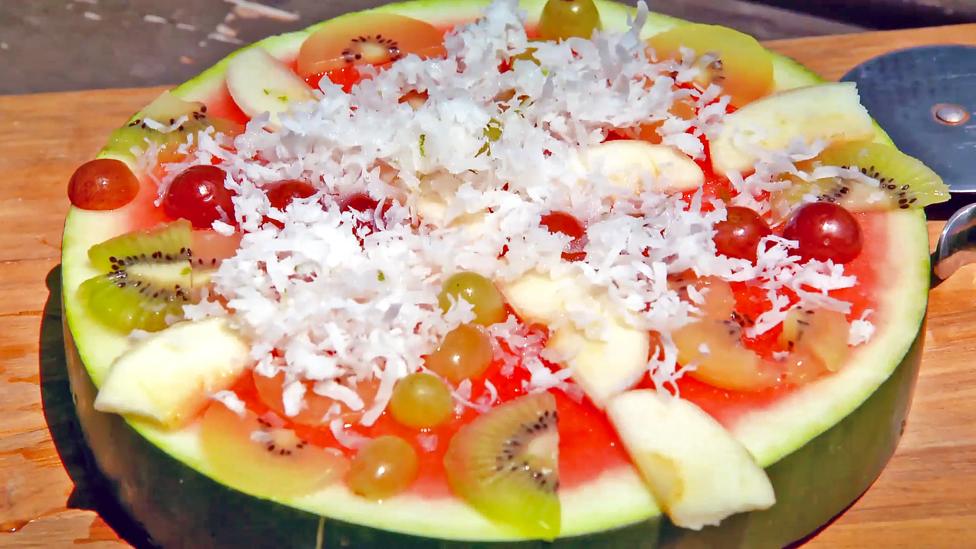 An even watermelon slice covered with apples, grapes, kiwi fruit and coconut shavings so that it looks like a pizza.