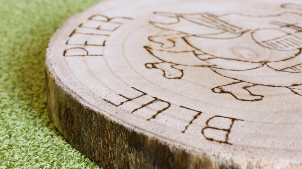 The edge of the log, showing the tree bark, and the word "Blue" carved into the woodgrain on the face side. Made by Macey from Bridgend, Wales.