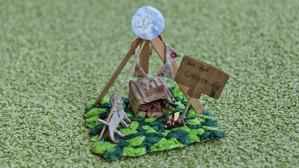 A mini model of the Blue Peter Garden, featuring green tissue paper for grass, a 'Bug Hotel' made from corrugated cardboad, a cardboard Henry dog and sign, and a tipi structure made from lolly sticks.