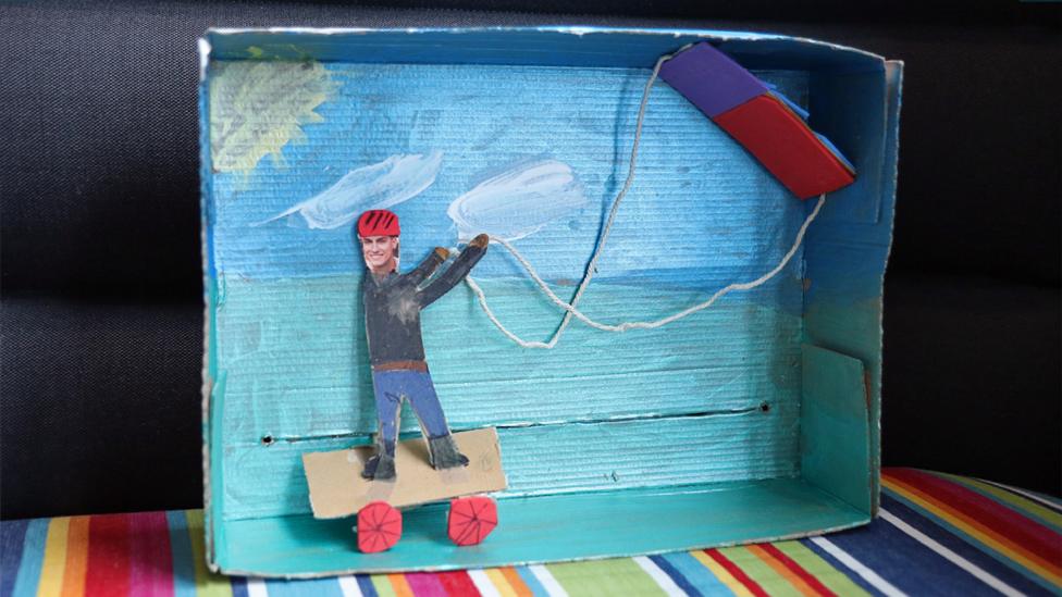A model of Richie on a landboard, holding a model kite, with two strings. In the background the sky, sun, clouds and grass have been painted.