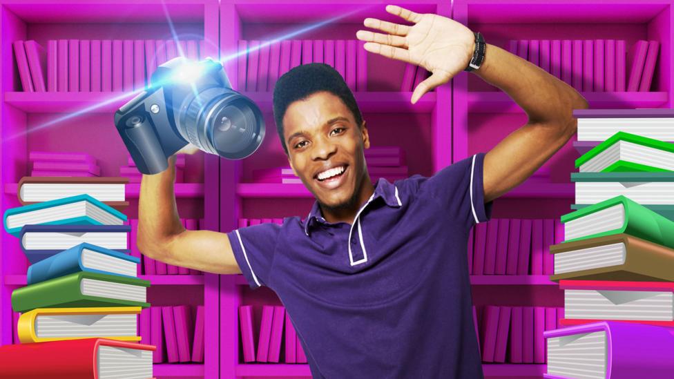 Rhys holding a camera surrounded by books and a book shelf.