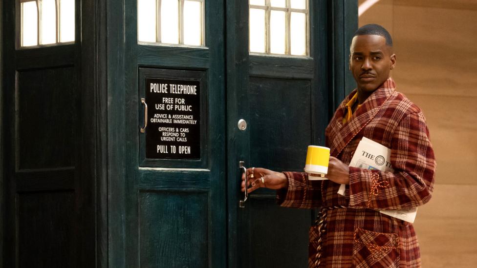 The Doctor, holding a newspaper and a cup of coffee, he is dressed in comfortable clothing and entering the TARDIS, the spaceship disguised as a blue police telephone box