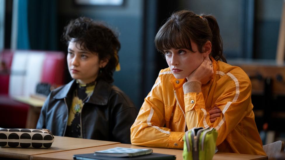 Jenna and Emily sit at a desk looking downcast