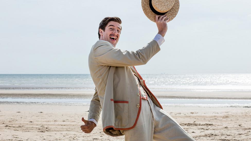 Billy Butlin holding his hat up and smiling on a beach