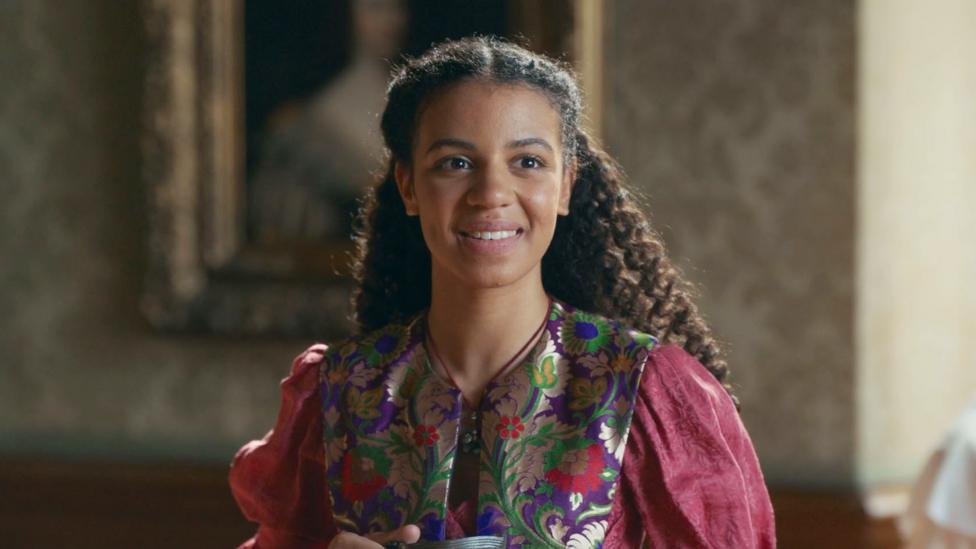 Ellie from The Lady Grace Mysteries smiles whilst wearing a light red dress.