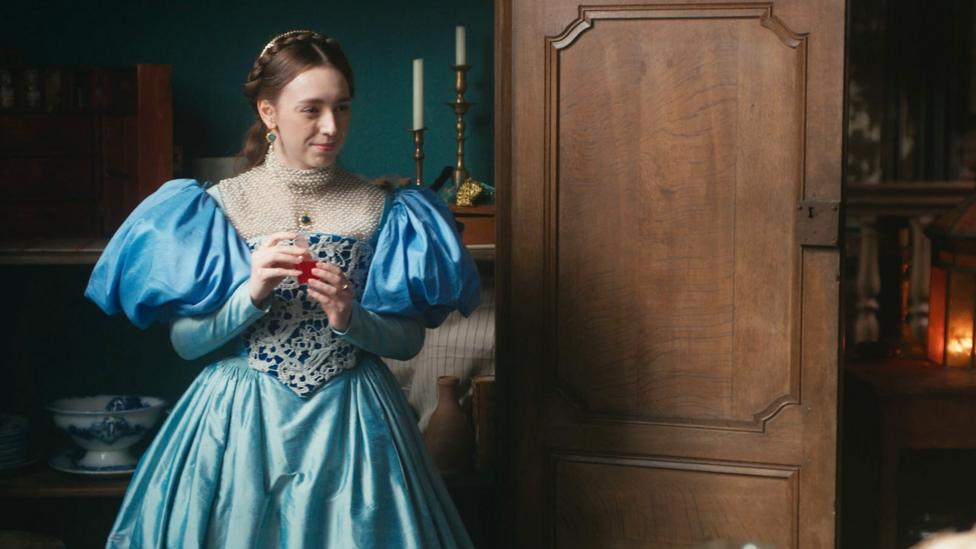 A young girl dressed in a blue Tudor-style dress waits by a large woodendoor.