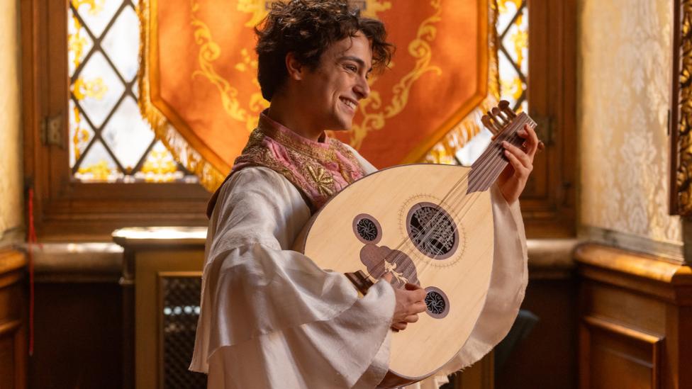 A man wears Tudor dress and carries an instrument.