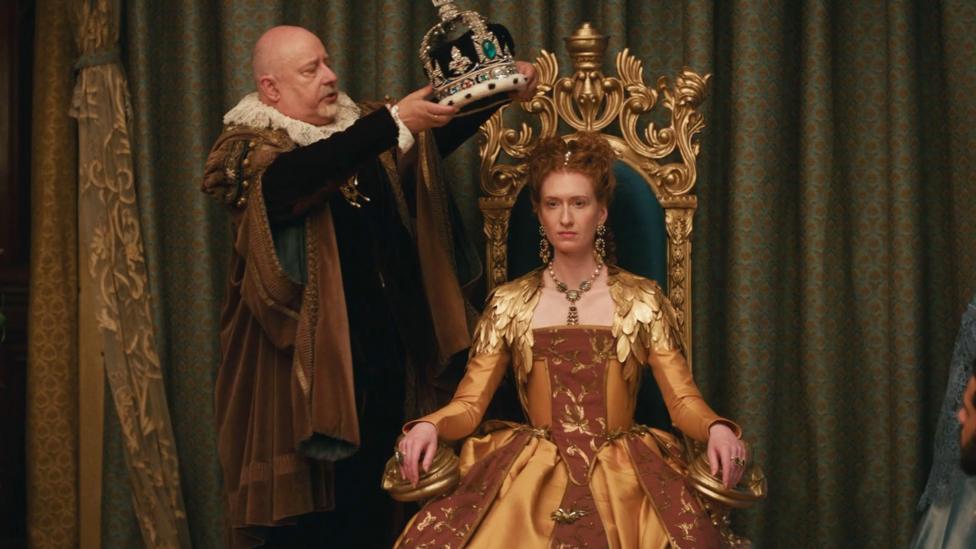 Image of a Queen sat on a throne with a man dressed in Tudor dress puts a crown on her head.
