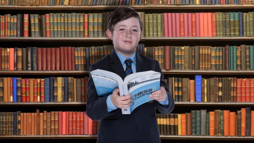 Maxwell, a character in So Awkward, stands holding a book in front of a library book shelf.