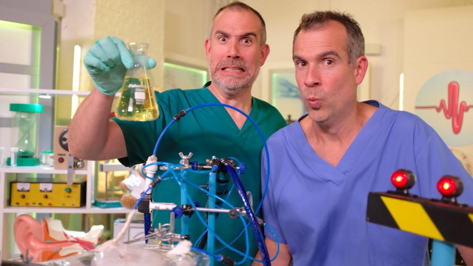 Dr Chris and Dr Xand stand in the lab looking shocked whilst holding a beaker of liquid.