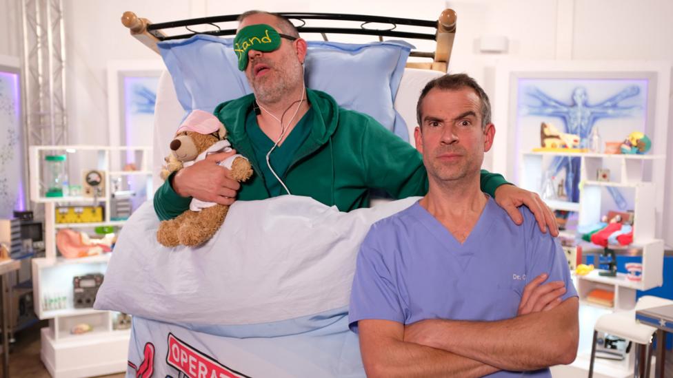 Image of Dr Chris with his arms crossed and Dr Xand asleep in bed behind him