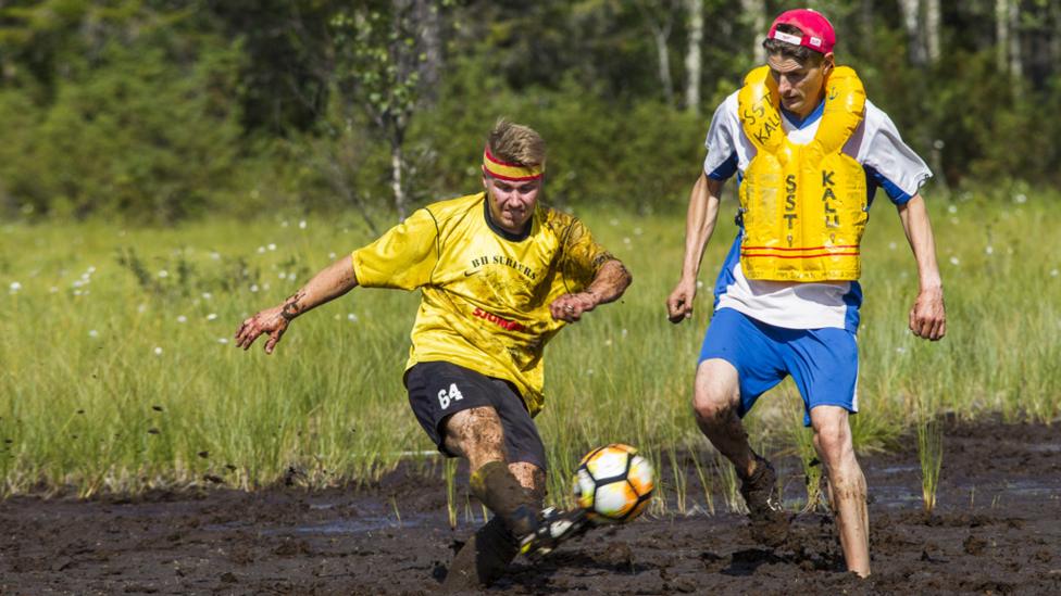 Two footballers covered in mud attempt to kick a football in a swamp.