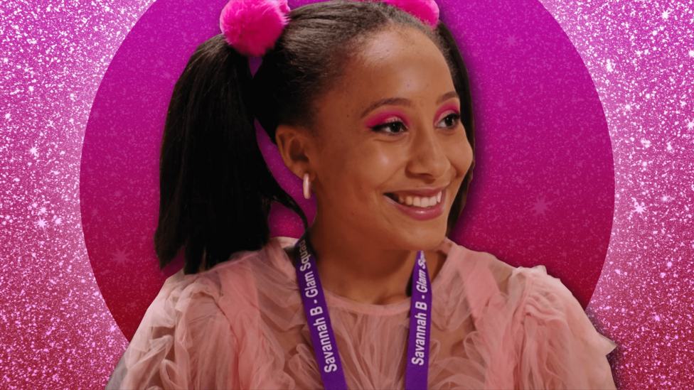 Candi-Rose is standing in front of a glittery pink background. She is wearing a pink lace blouse and has bright pink eyeshadow