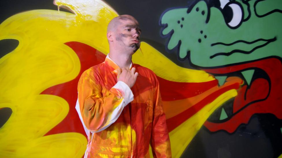 Gav in a boiler suit next to a dragon picture.