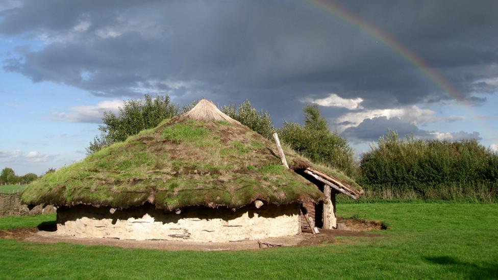 A Bronze Age roundhouse in a field with a rainbow cutting through the dark clouds in the background