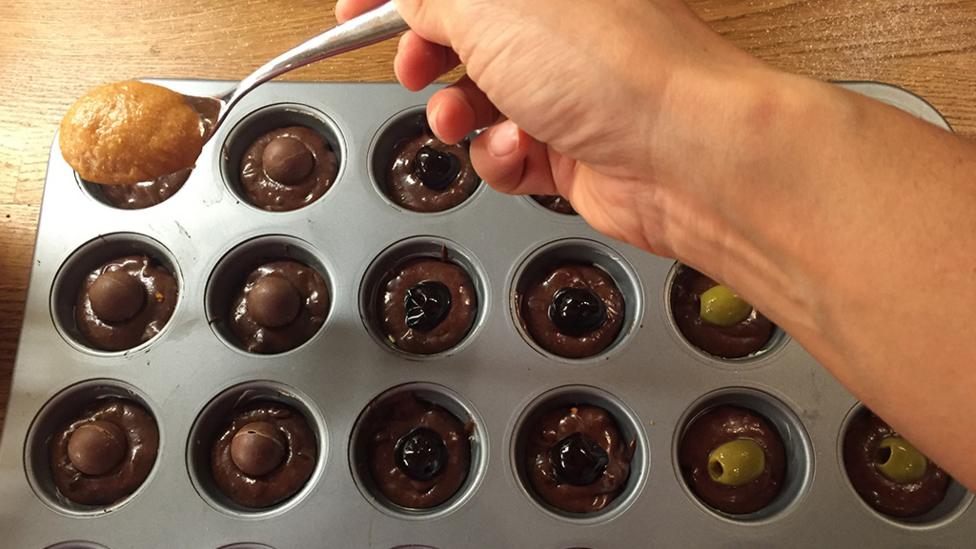 Spooning pumpkin batter onto cupcakes with chocolate batter and olives or cherries inside.