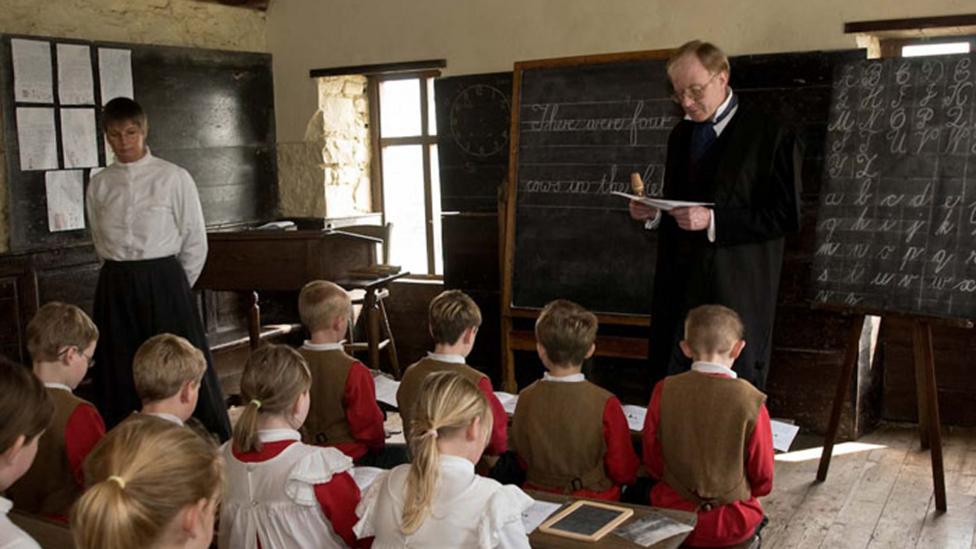 A school teacher dressed in a Victorian outfit teaching school children in an old classroom with a blackboard and desks