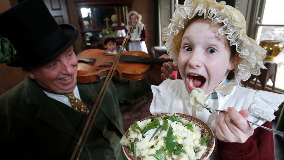 A man playing the fiddle and a young girl eating, both costumed in Victorian garb