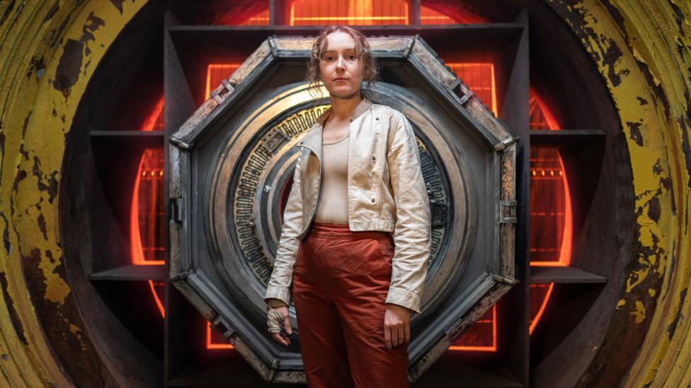 Rose Ayling-Eliss as Aliss in Doctor Who, she is a young woman wearing a white jacket and red cargo trousers.
