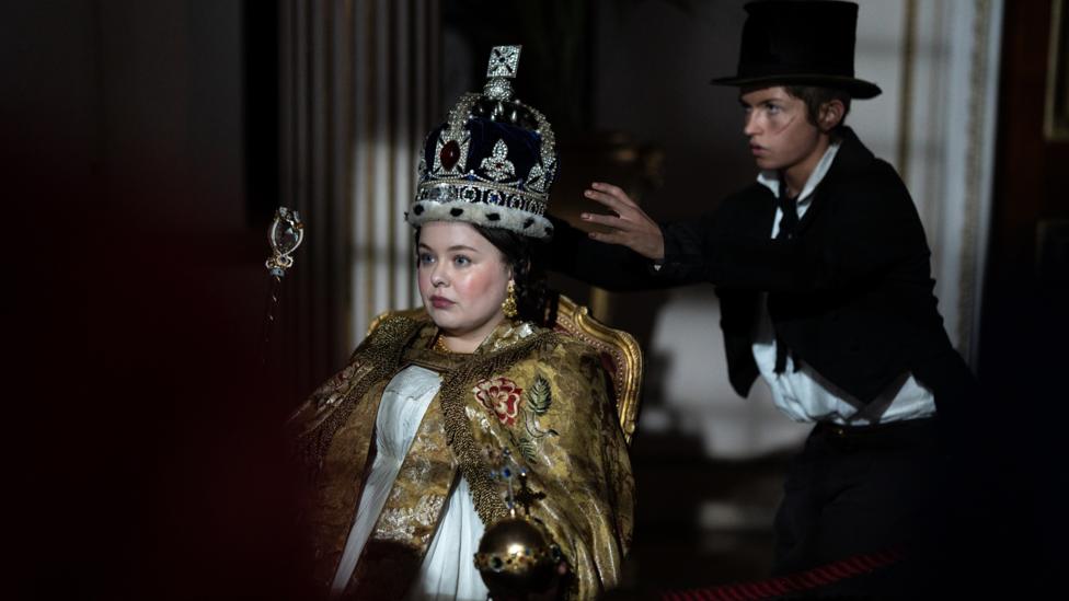 Dodger creeps up behind Queen Victoria, his hands moving towards the crown on her head.