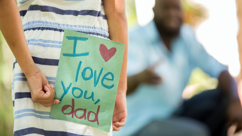 A girl holding a father's day card behind her back, walking towards her dad.