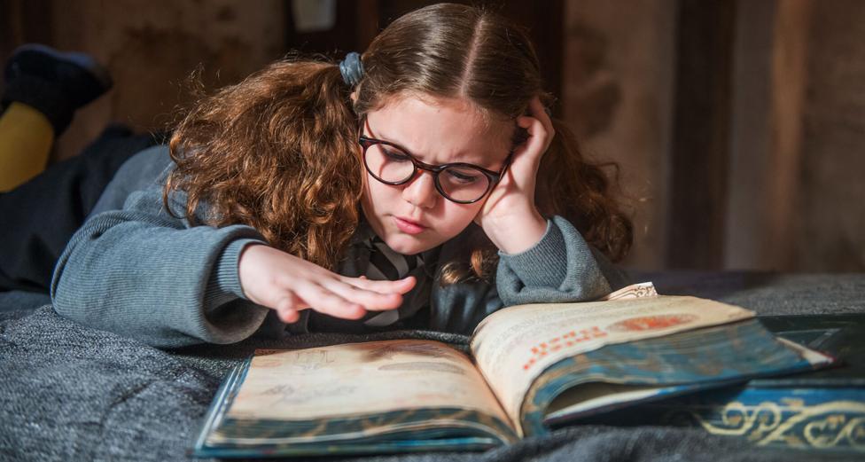 A girl lay on top of a bed with a large old book and hovers her hand over the page as she reads, (Maud).