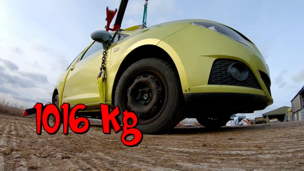 A car is connected to a crane, text says that the car weighs 1016 kilograms.