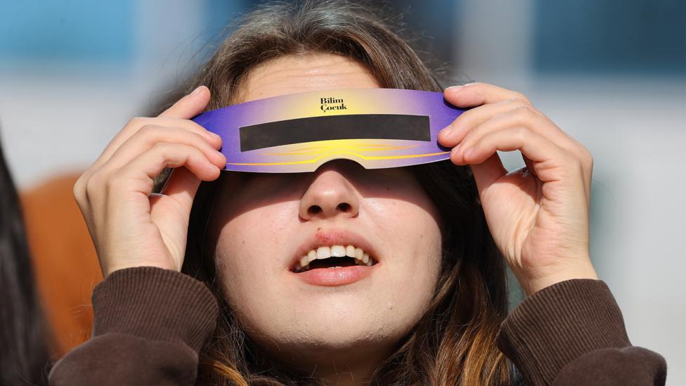 A person looks up in wonder at an eclipse, she is wearing specialised solar eclipse safety glasses.
