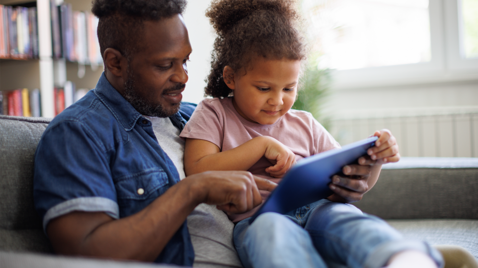 A father sat on a sofa with his daughter and they are both looking at a computer tablettablet
