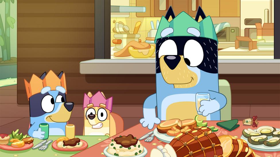 Bluey, Bingo and Dad sit at the Christmas table with food in front of them