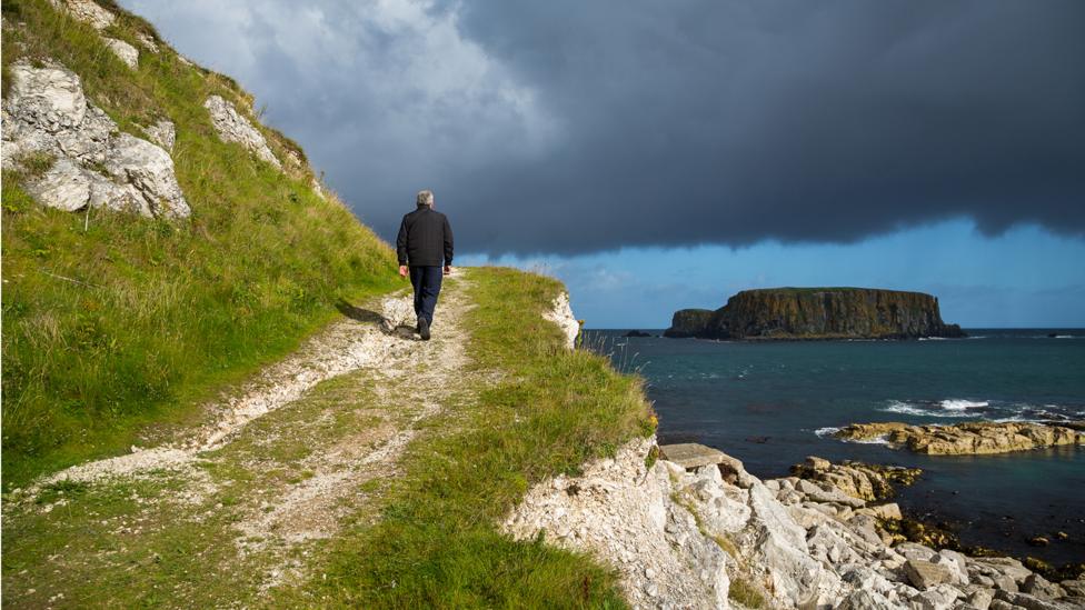 Image of a man walking along a hillside path, with the sea on the right below him and the Giants Causeway.