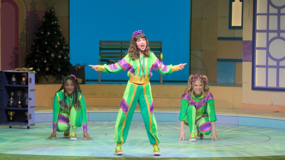 Darcy Dribble from the CBeebies Christmas Panto 2025: Cinderella, dances confidently between her two other sisters, who are crouched low as through they are about to run forwards.